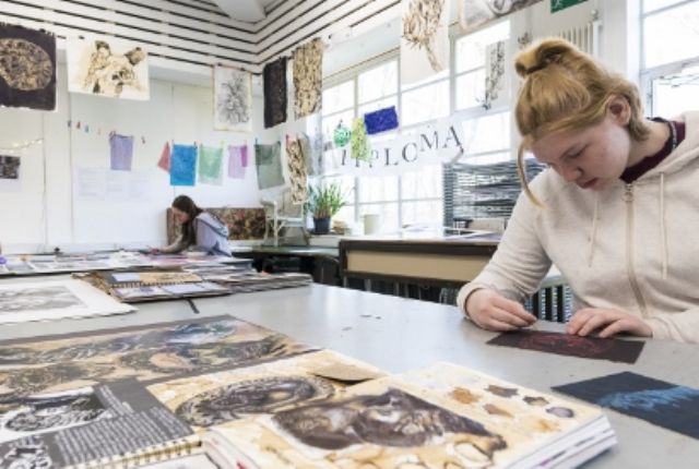 student drawing on black paper with red pen, surrounded by artwork portfolios on the desks